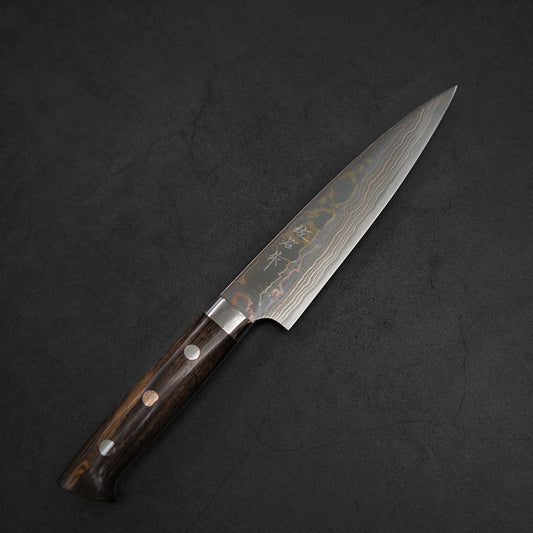 The Takeshi Saji rainbow damascus aogami#2 petty knife 150mm features a wave-patterned damascus steel blade with engraved characters and a dark wooden handle, shown resting on a black textured surface.