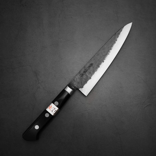 The Teruyasu Fujiwara Denka petty knife 150mm, featuring a Damascus-patterned blade with Japanese characters and a black handle marked X2, lies on a dark gray textured surface.