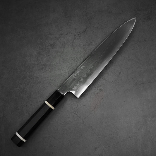 A Yoshikazu Ikeda shirogami#2 honyaki gyuto 240mm with a dark handle and sharp, polished blade rests on a gray textured surface. Subtle etched patterns grace the Hitachi shirogami#2 steel blade near the spine.