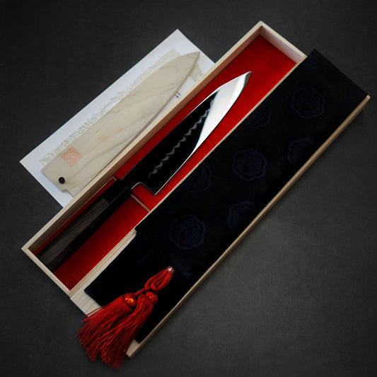 A Yoshikazu Ikeda shirogami #1 honyaki kiritsuke gyuto 240mm with a patterned blade rests in an open wooden box lined with red fabric, accompanied by a wooden sheath, black cloth cover with red tassel, and certificate.