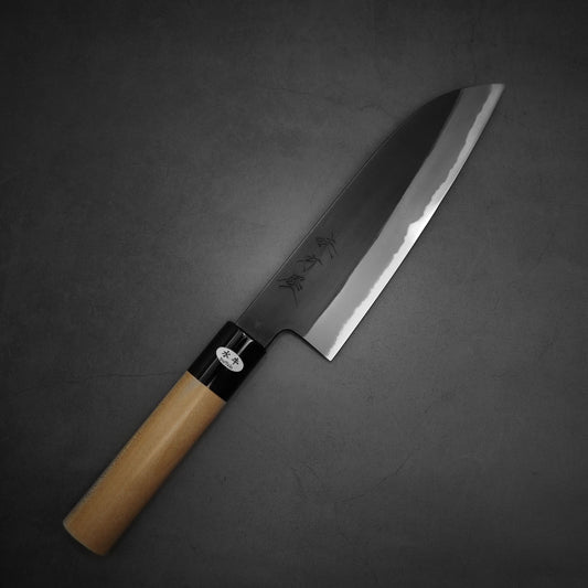The Hinoura kurouchi shirogami #2 170mm santoku, with a wooden handle and wide dark blade engraved with characters, is elegantly placed on a dark grey surface.
