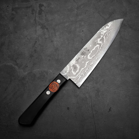 The Shigeki Tanaka VG10 damascus santoku 165mm, featuring a dark handle and patterned blade, rests on a dark textured surface—a striking example of Shigeki Tanaka’s craftsmanship.