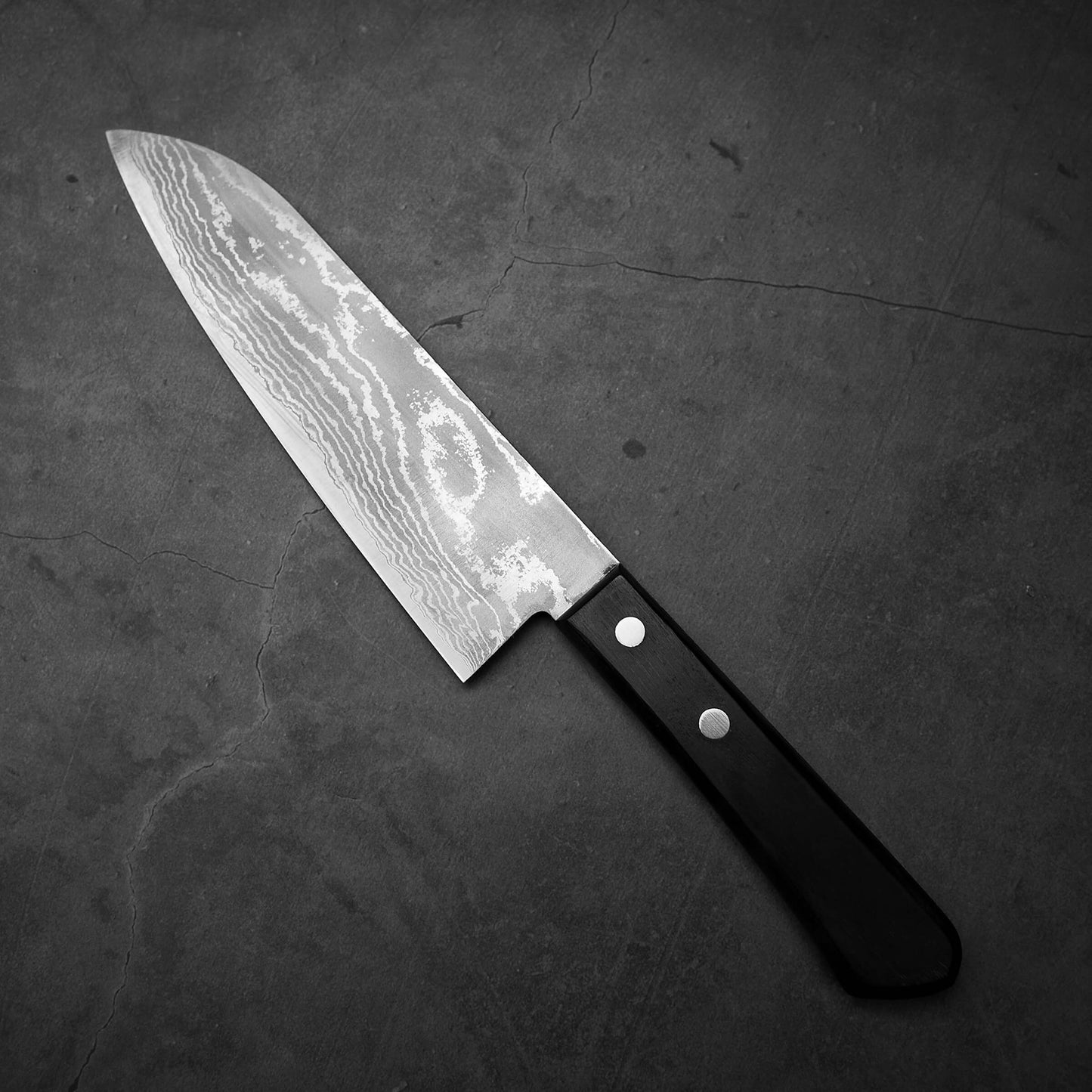 The Shigeki Tanaka VG10 damascus santoku 165mm features a patterned VG10 stainless steel blade and black handle with two metal rivets, embodying Shigeki Tanaka’s tradition of hand-crafted knife-making. It rests on a dark, cracked stone surface.
