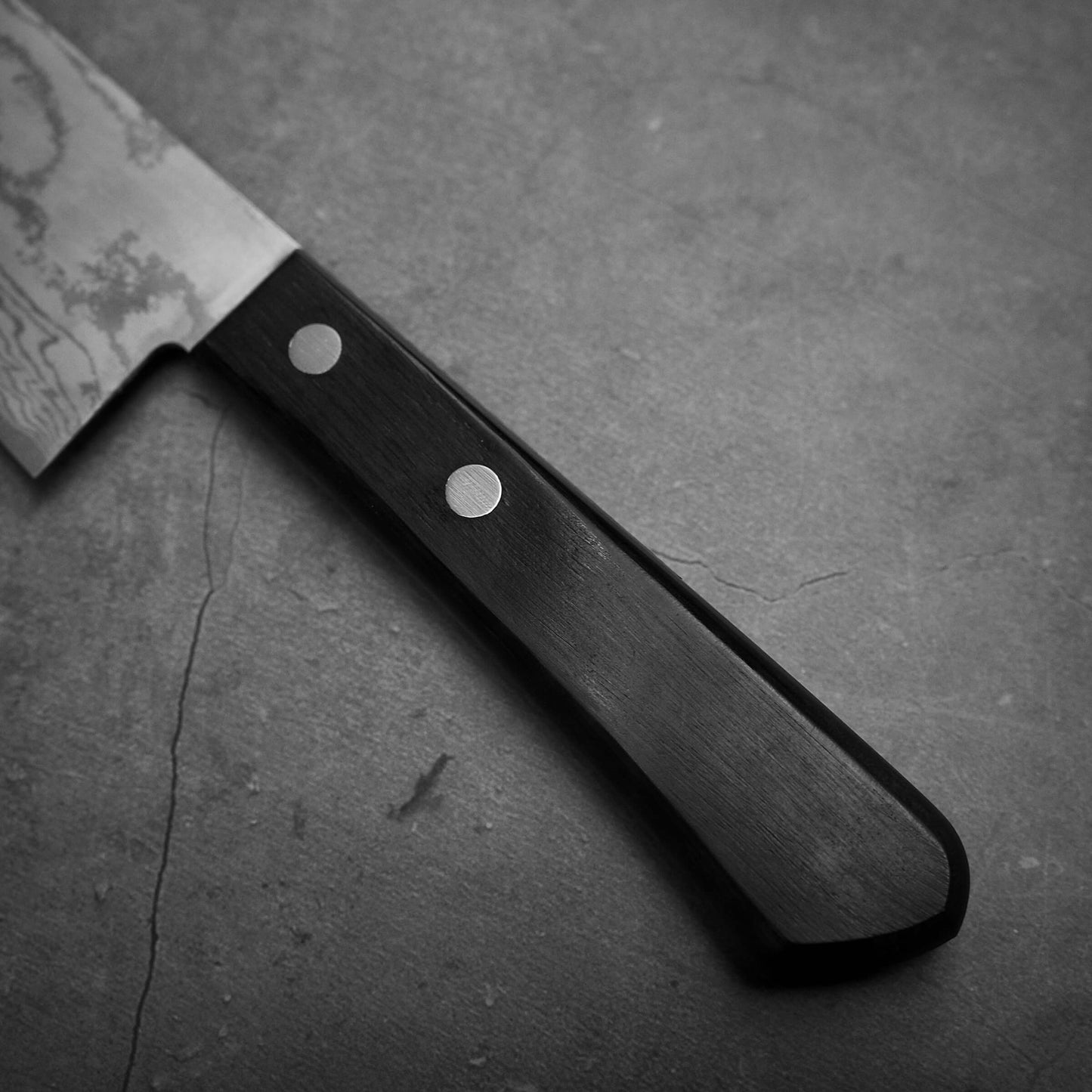 A close-up of the Shigeki Tanaka VG10 damascus santoku 165mm knife with a dark wooden handle and two rivets, featuring a VG10 stainless steel blade, lying on a textured, cracked gray surface.