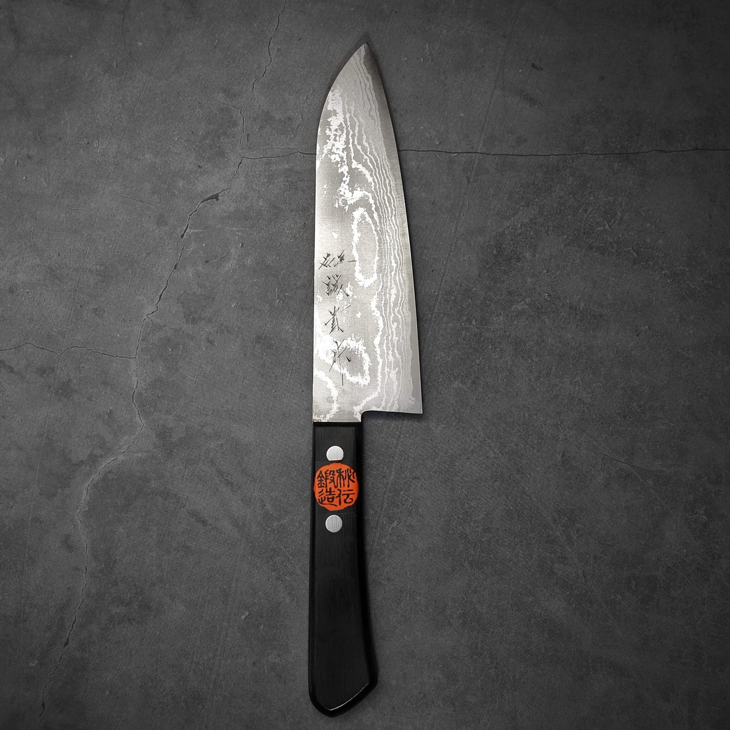 The Shigeki Tanaka VG10 damascus santoku 165mm, crafted by Shigeki Tanaka, features a black handle with two metal rivets and a red sticker with Japanese characters, plus a wavy blade pattern. It rests on a dark gray textured surface.