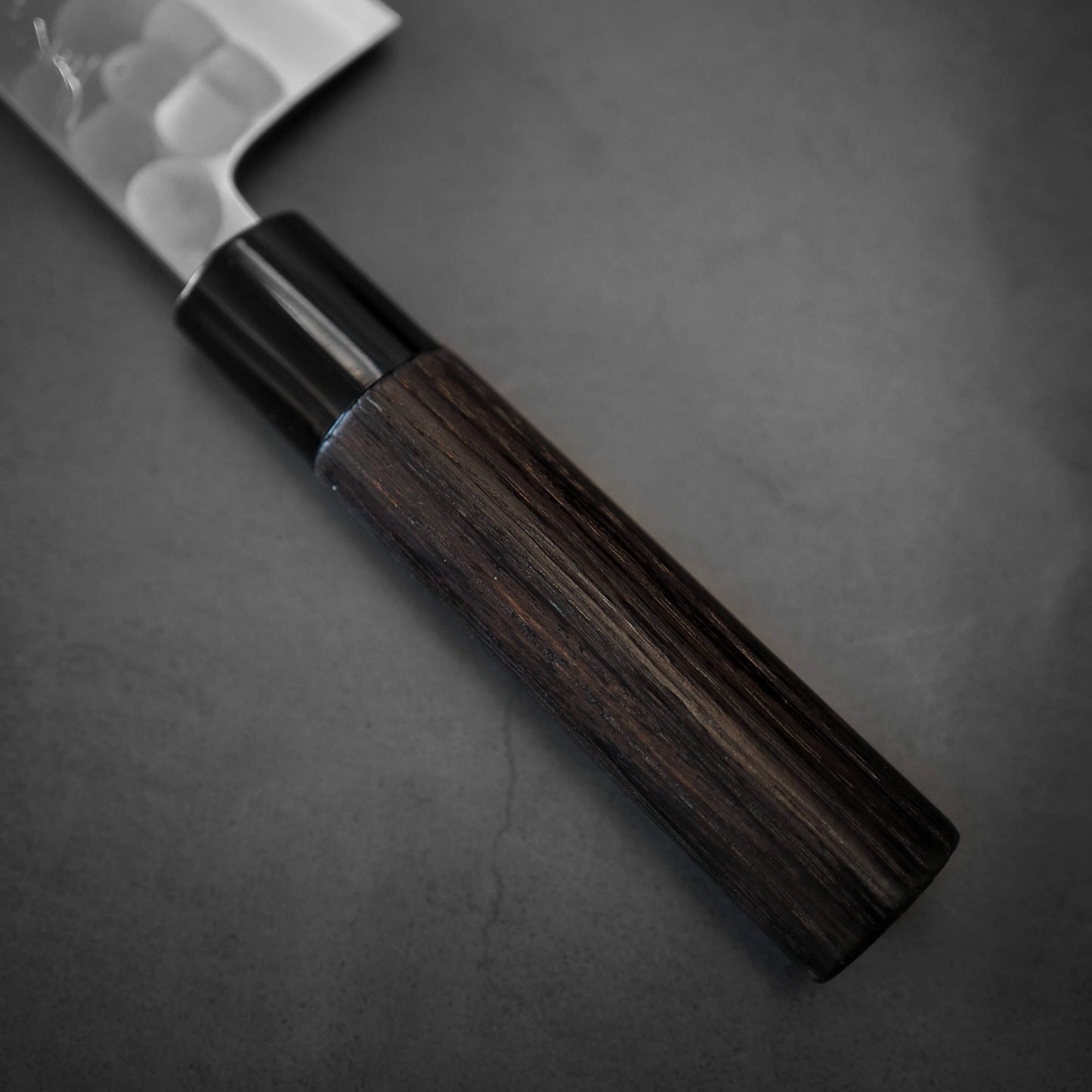 A close-up of the Yoshikane tsuchime SKD santoku 165mm knife by Yoshikane, featuring a dark wooden handle and an SKD steel blade, placed on a dark gray surface.