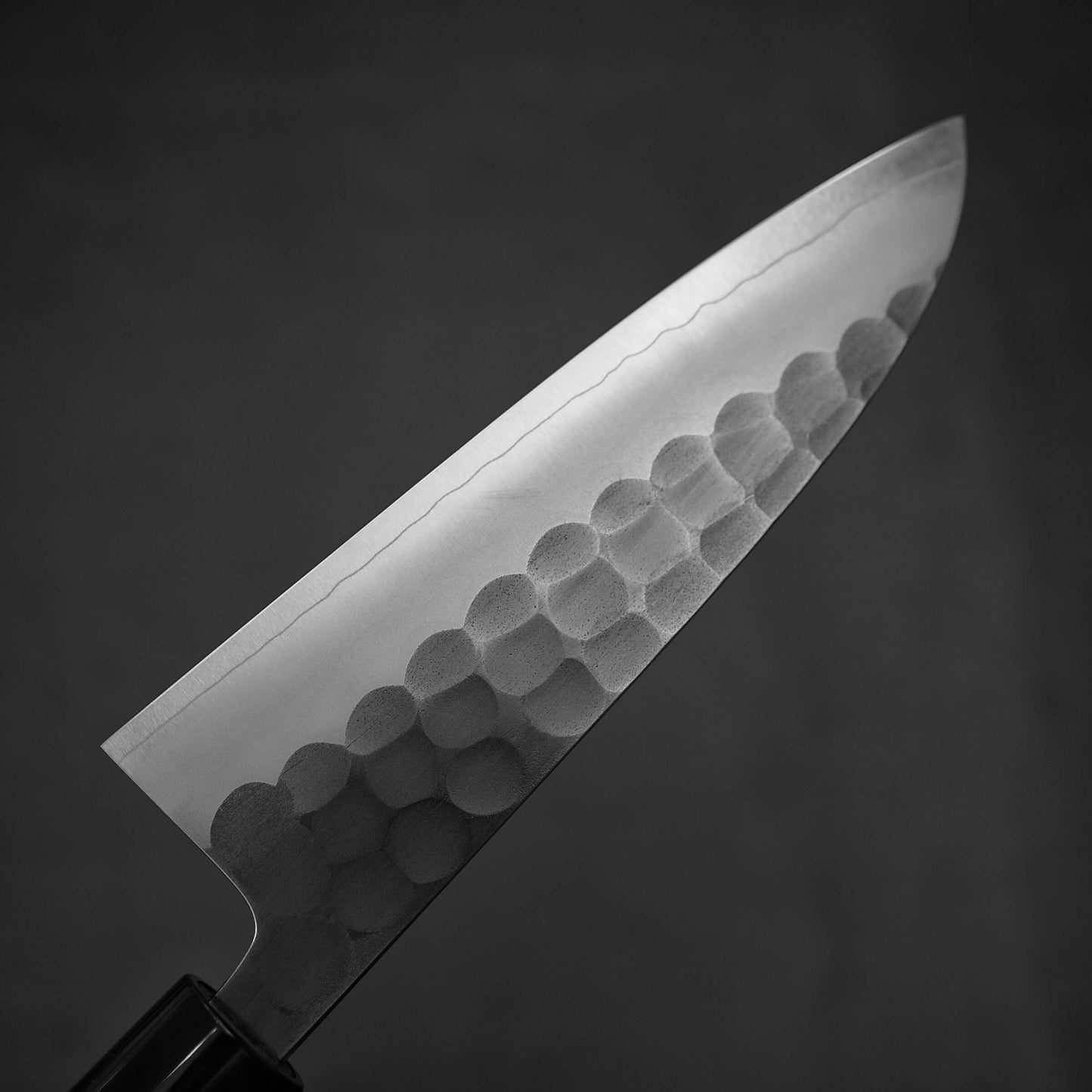 A close-up of the Yoshikane tsuchime SKD santoku 165mm knife blade shows its hammered SKD steel lower half and smooth, sharp edge, set against a dark, blurred background.