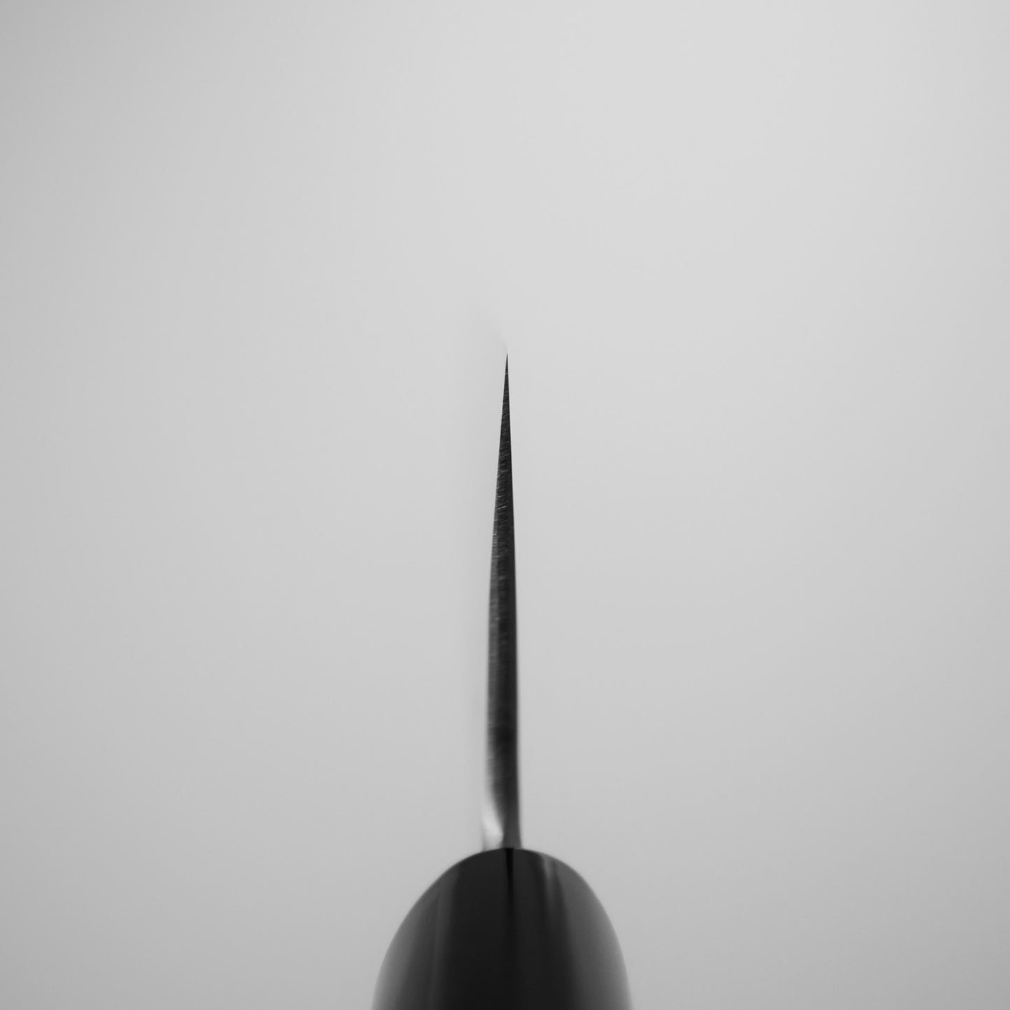 Close-up black-and-white photo of the sharp, thin tip of the Yoshikane tsuchime SKD santoku 165mm knife by Yoshikane, seen from above against a plain light background.