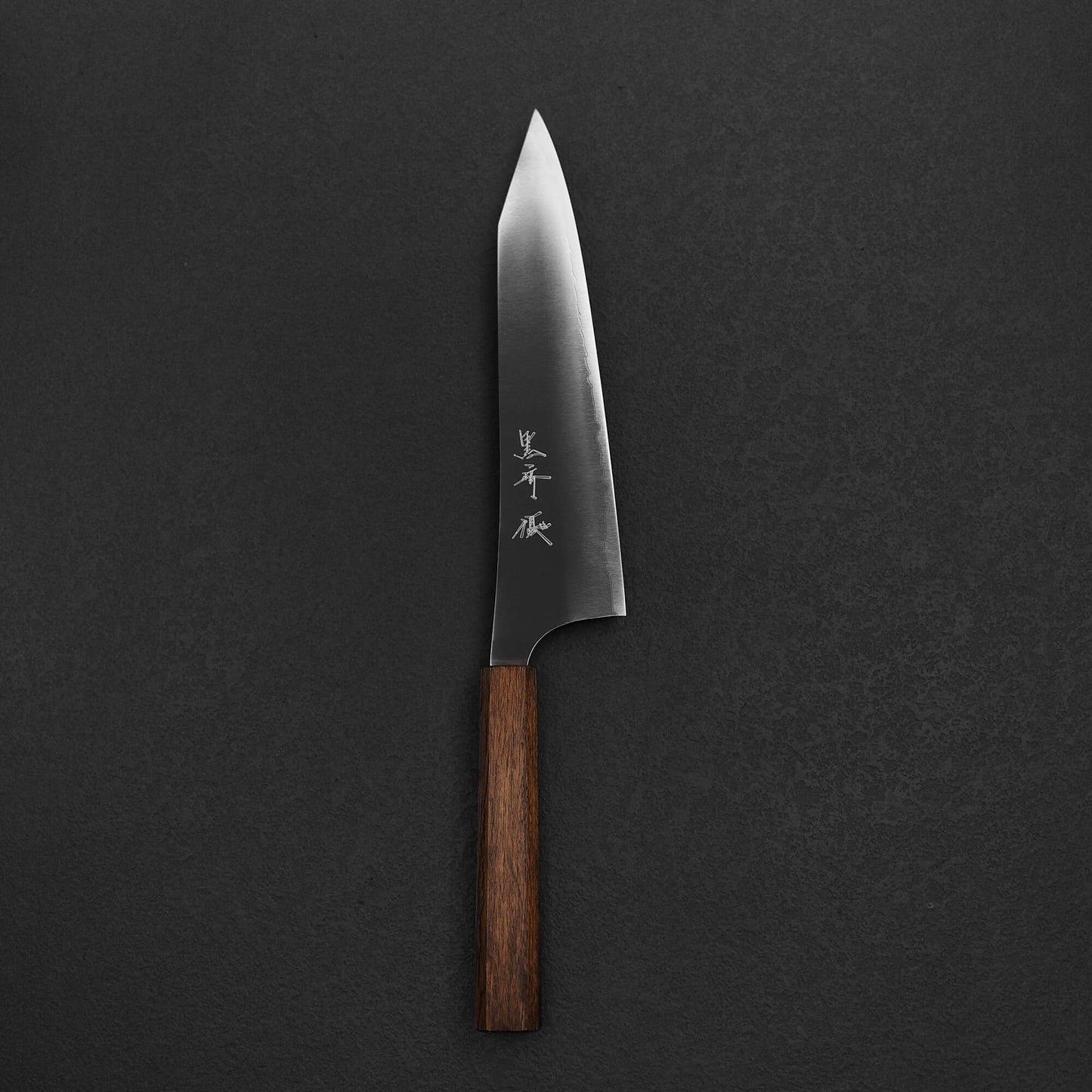 The Yu Kurosaki HAP40 Gekko kiritsuke gyuto 210mm chef’s knife features a dark wooden handle and a polished HAP40 steel blade with Japanese engraving, shown resting on a dark textured surface.