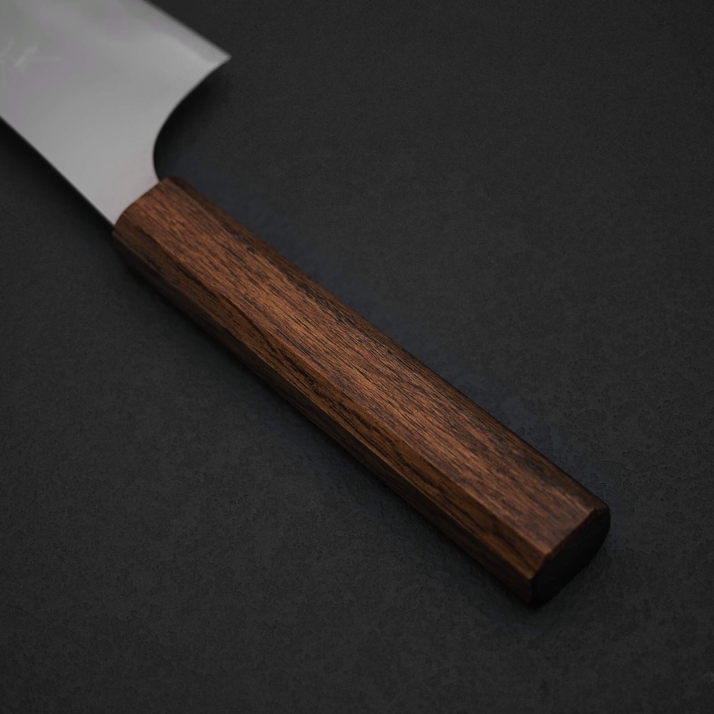 A close-up of the Yu Kurosaki HAP40 Gekko kiritsuke gyuto 210mm, with a wooden handle and HAP40 steel blade, resting on a dark surface. Only the handle and part of the blade from this Yu Kurosaki knife are visible.