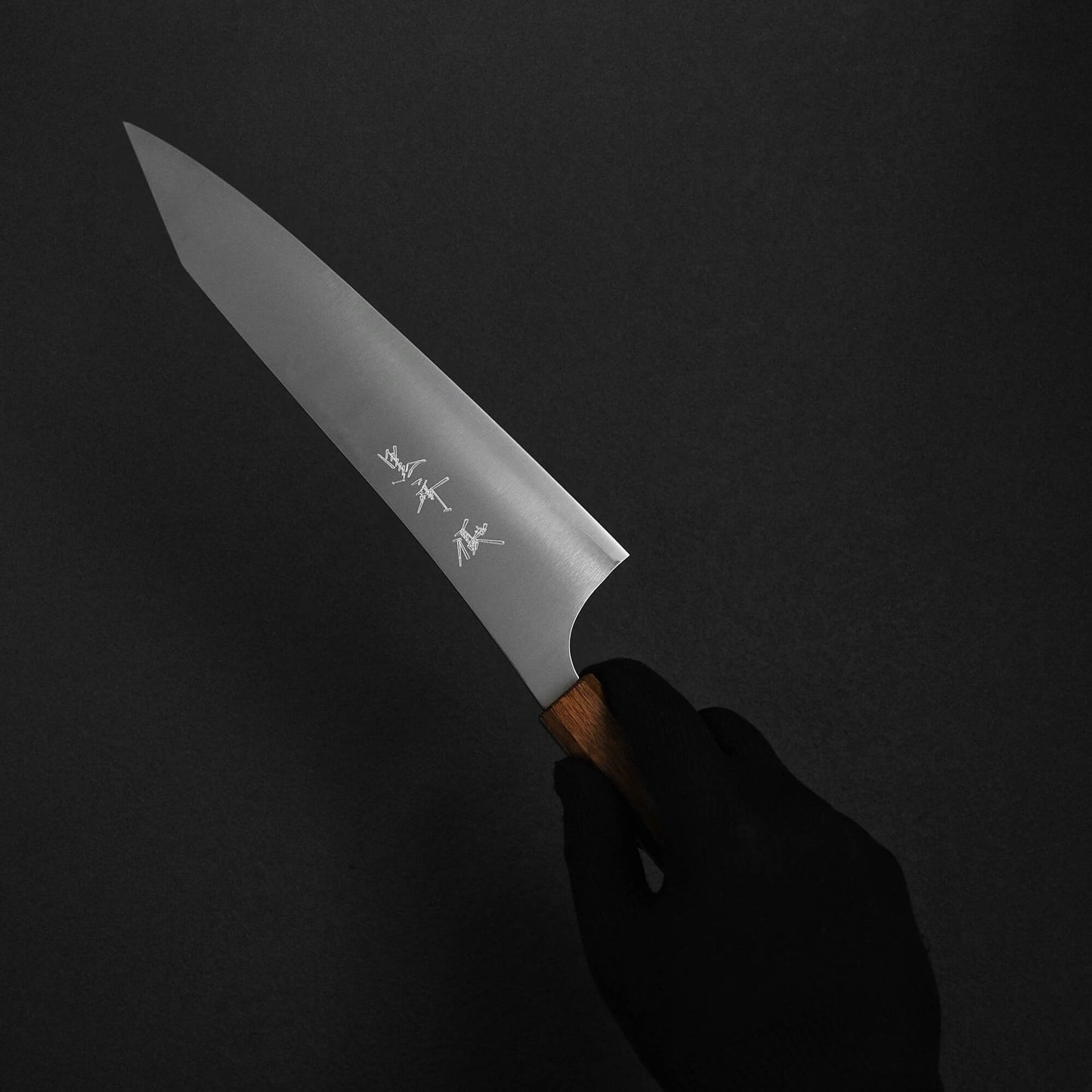 A hand in a black glove holds the Yu Kurosaki HAP40 Gekko kiritsuke gyuto 210mm, featuring a wooden handle and engraved Japanese characters on its HAP40 steel blade, set against a dark background.