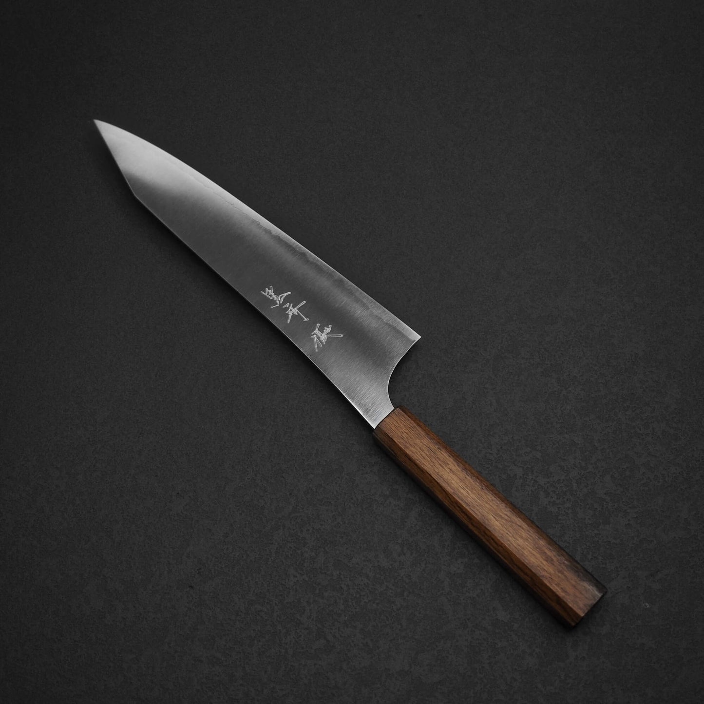A Yu Kurosaki HAP40 Gekko kiritsuke gyuto 210mm chef’s knife with a wooden handle and glossy, sharp HAP40 steel blade adorned with kanji sits on a dark, textured surface.