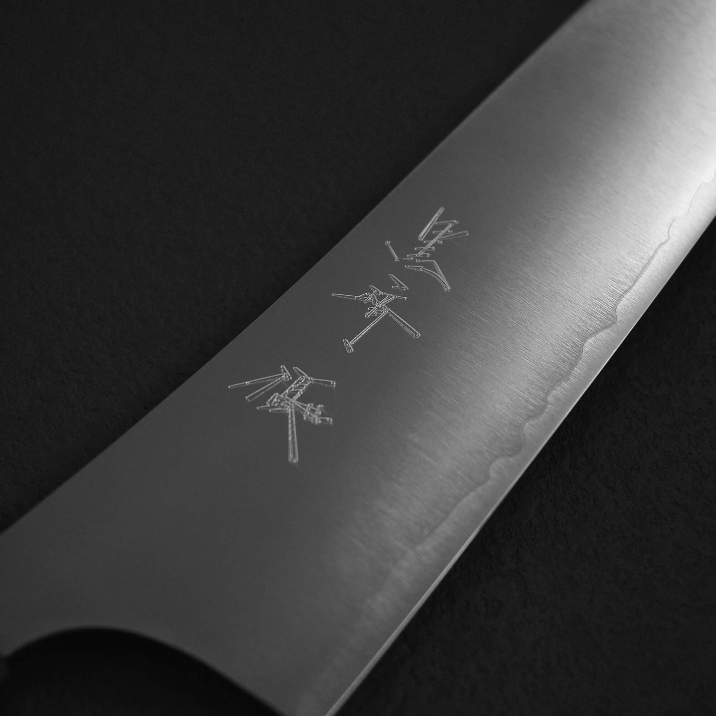 A close-up of the Yu Kurosaki HAP40 Gekko kiritsuke gyuto 210mm blade, featuring engraved kanji and a wavy edge pattern, set against a dark background.