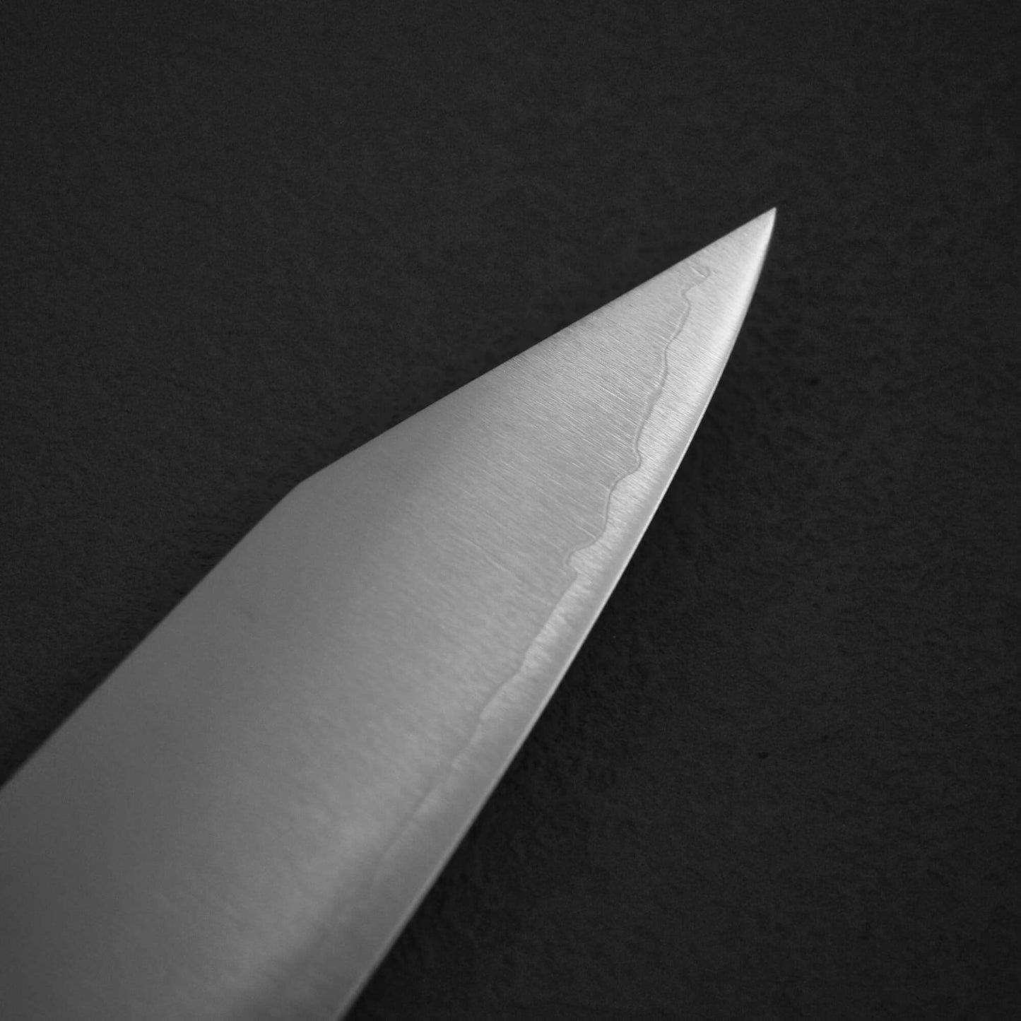 A close-up of the pointed tip of the Yu Kurosaki HAP40 Gekko kiritsuke gyuto 210mm blade, featuring a distinct wavy pattern near the edge, set against a dark textured background.