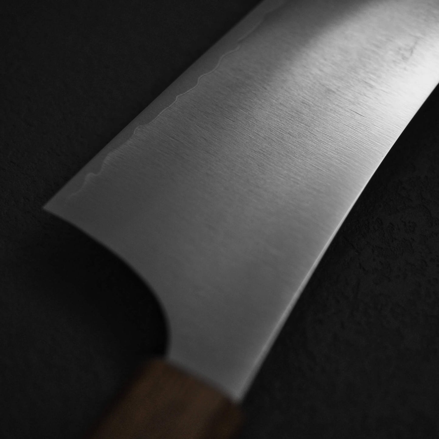Close-up of the Yu Kurosaki HAP40 Gekko kiritsuke gyuto 210mm features its unique wavy blade pattern and wooden handle, set against a dark textured background.