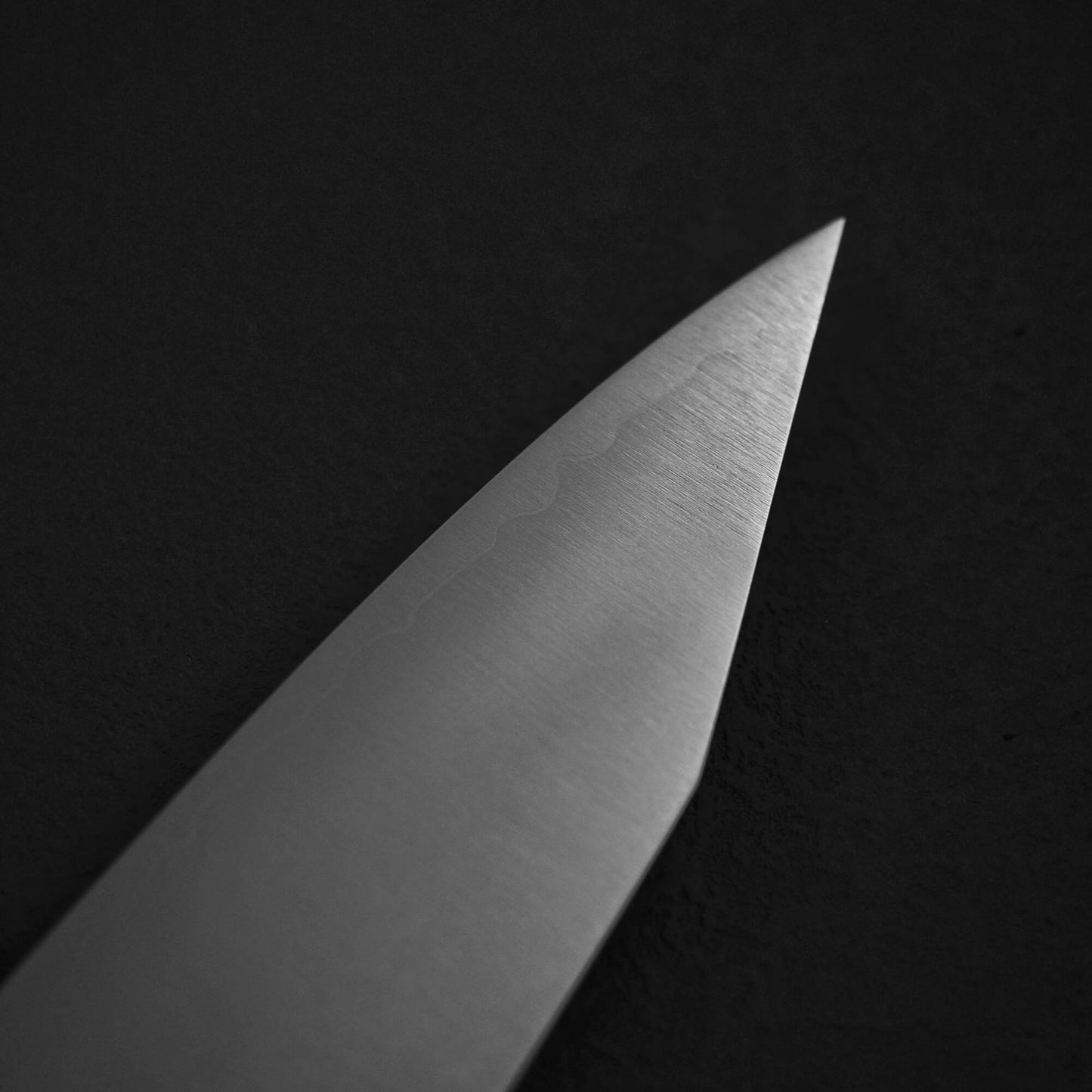 A close-up of the pointed tip of the Yu Kurosaki HAP40 Gekko kiritsuke gyuto 210mm knife, crafted by Yu Kurosaki from HAP40 steel, set against a dark, textured background.