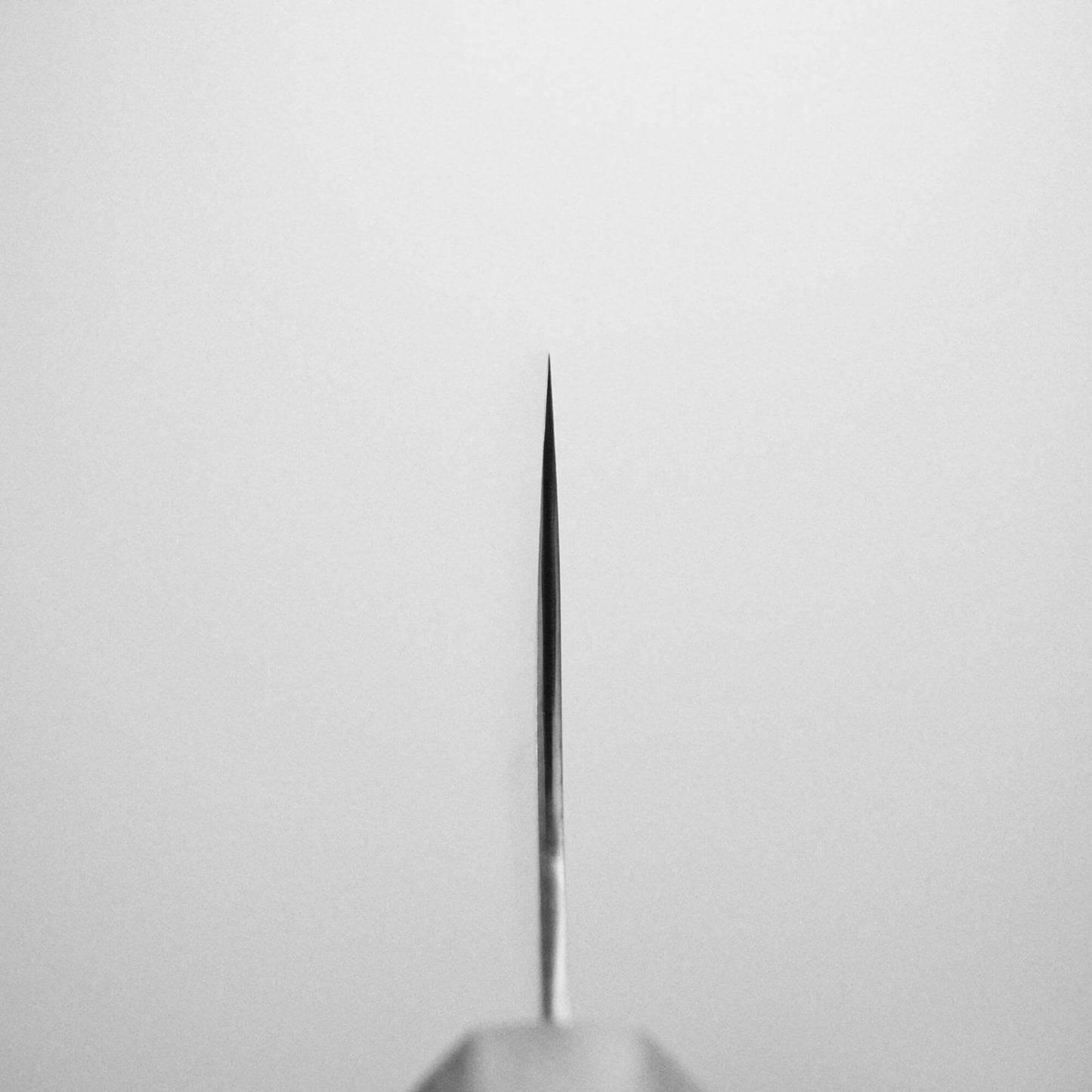 Centered close-up of the Yu Kurosaki HAP40 Gekko kiritsuke gyuto 210mm knife tip pointing upward against a light gray background, highlighting the thin, symmetrical HAP40 steel blade with its sharp tip in clear focus.