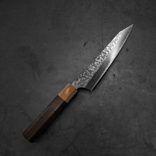 A Japanese-style kitchen knife with a hammered steel blade and a wooden handle, lying on a dark, textured concrete surface.