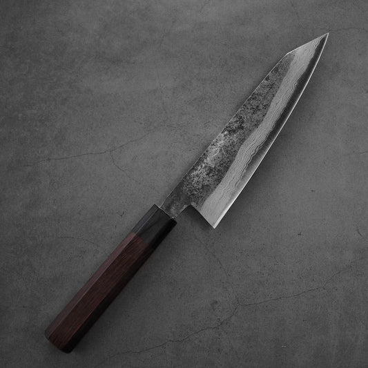 A Shigeki Tanaka kurouchi damascus aogami#2 kiritsuke gyuto 190mm chefs knife with a dark wooden handle and patterned blade rests on a gray, cracked surface.