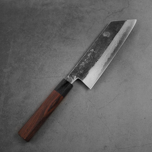 The Shigeki Tanaka kurouchi damascus aogami#2 kiritsuke nakiri 170mm, featuring a dark wooden handle and patterned blade, rests on a gray stone surface.