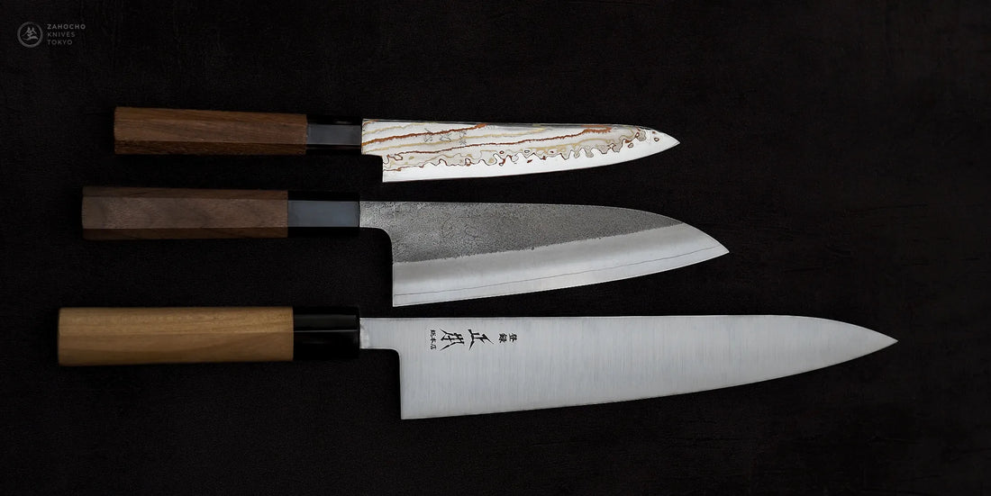 Three Japanese kitchen knives on black background: Hatsukokoro rainbow damascus petty knife, Yoshikane santoku, and Masamoto KS gyuto chef's knife. All feature wooden handles.