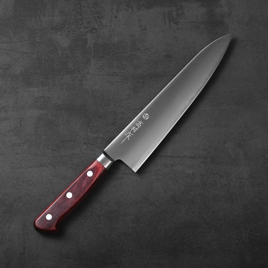 A Takamura Japanese chef’s knife with a satin finish, stainless steel blade and a polished, red wooden handle rests on a textured black surface.