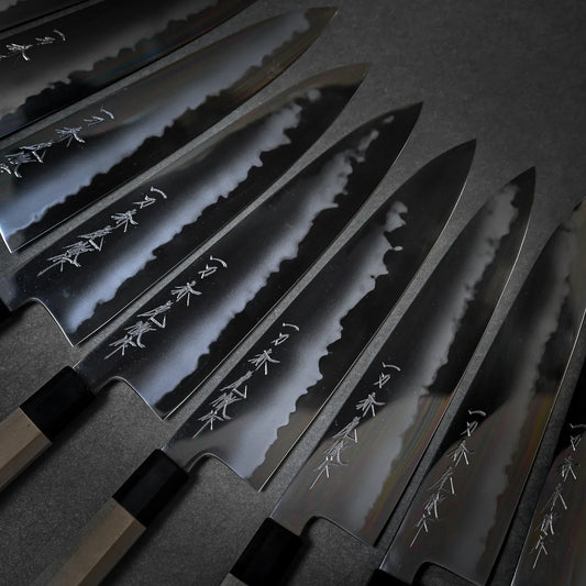 Collection of Tanaka gyuto knives arranged diagonally on grey background showing kanji and the contrast between cladding and core steel