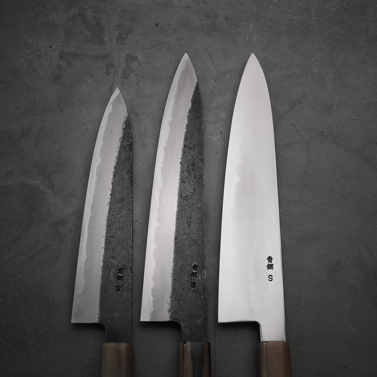 Three large Yoshikazu Tanaka kurouchi AS gyuto 210mm knives are arranged side by side on a dark textured surface; the two on the left have a rustic finish, while the polished knife on the right highlights Yoshikazu Tanakas signature craftsmanship.