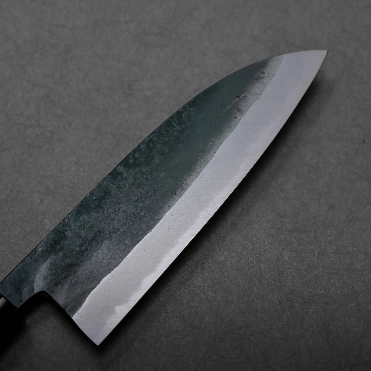 A close-up of a dark-handled Japanese knife, showing part of the reflective, hand-forged blade in detail.