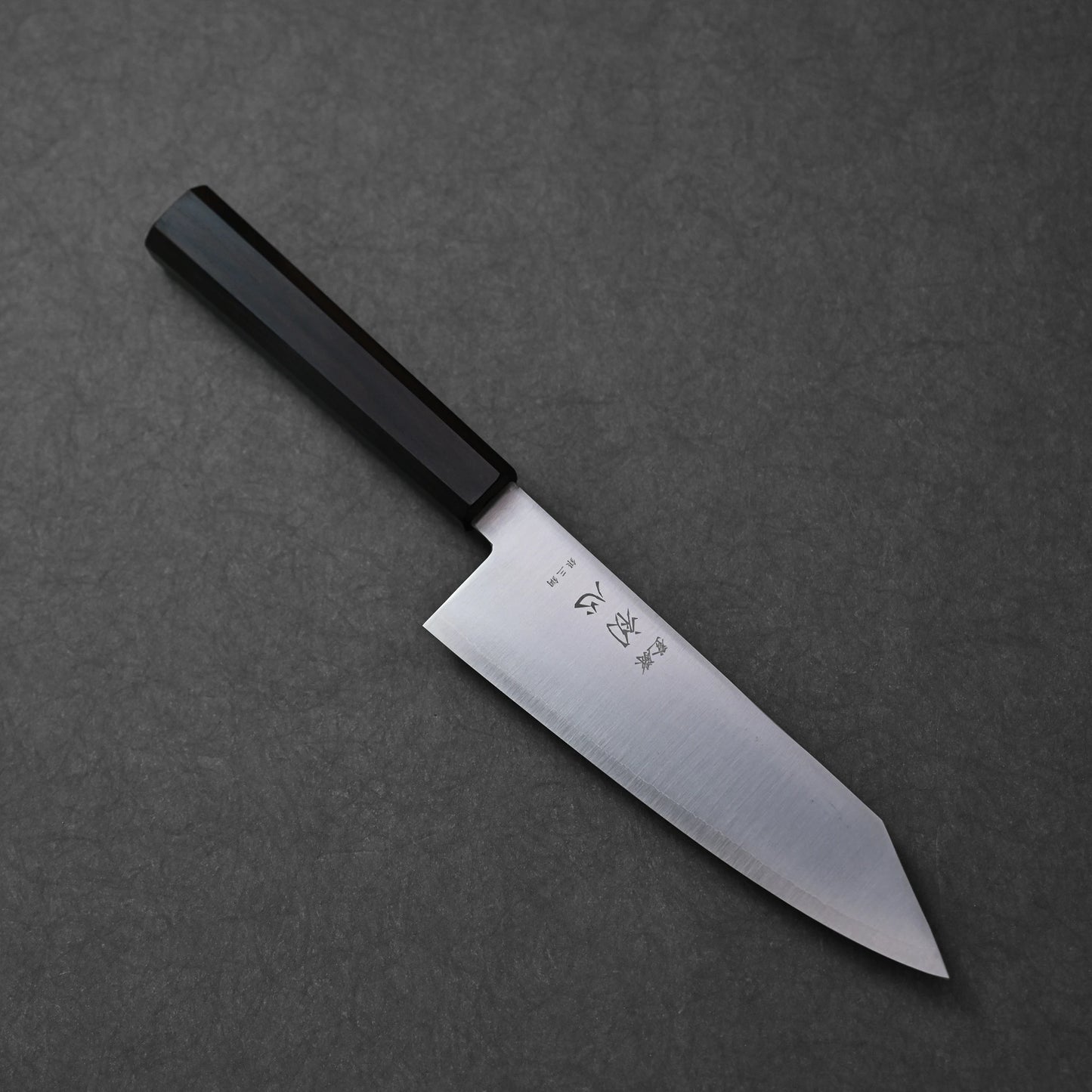 Bunka knife made with ginsan stainless steel; photographed in angled orientation to showcase the blade profile, pointed tip, engraved kanji near the handle, and the deep finish of the dark octagonal ebony handle.