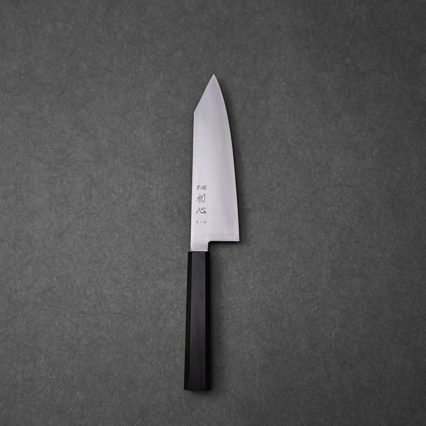 Bunka knife made with ginsan stainless steel; photographed to showcase the full blade profile, pointed tip, vertically engraved kanji near the handle, and the dark octagonal ebony handle, all set against a textured grey surface.