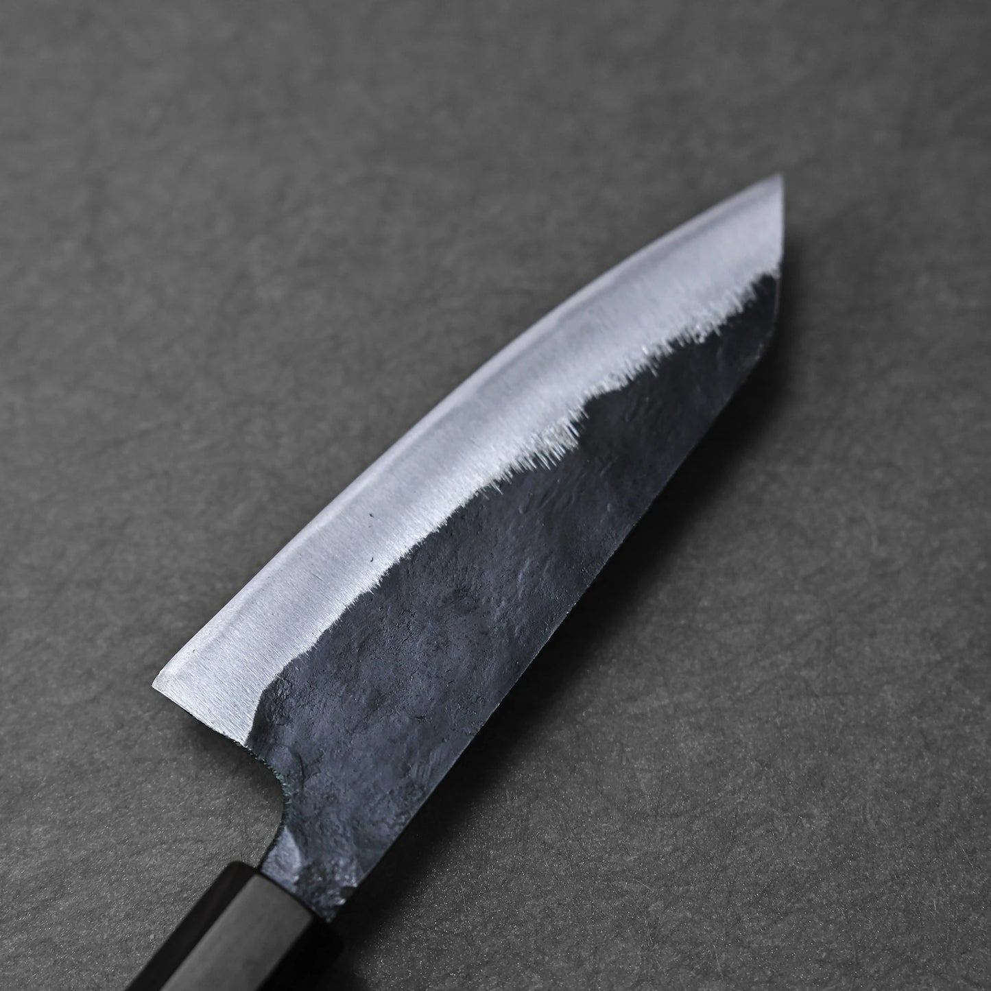 Close-up of the Hatsukokoro Kurouchi Aogami#2 Ko-Bunka 120mm kitchen knife by Hatsukokoro, featuring a black handle and a partially polished Aogami #2 blade on a dark matte surface.