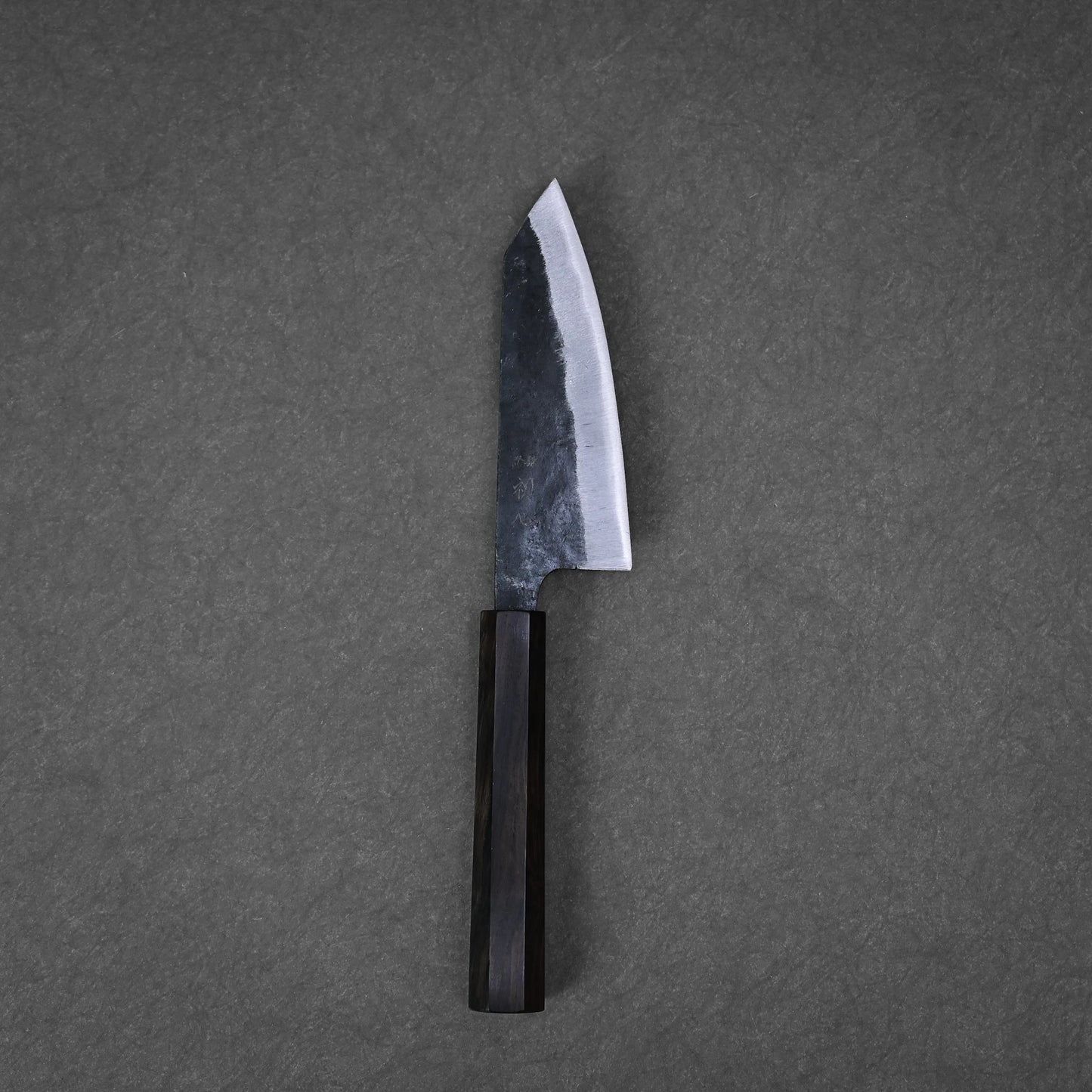A Hatsukokoro kurouchi aogami#2 ko-bunka 120mm kitchen knife with a dark wooden handle and wide, slightly curved blade rests on a textured gray surface.