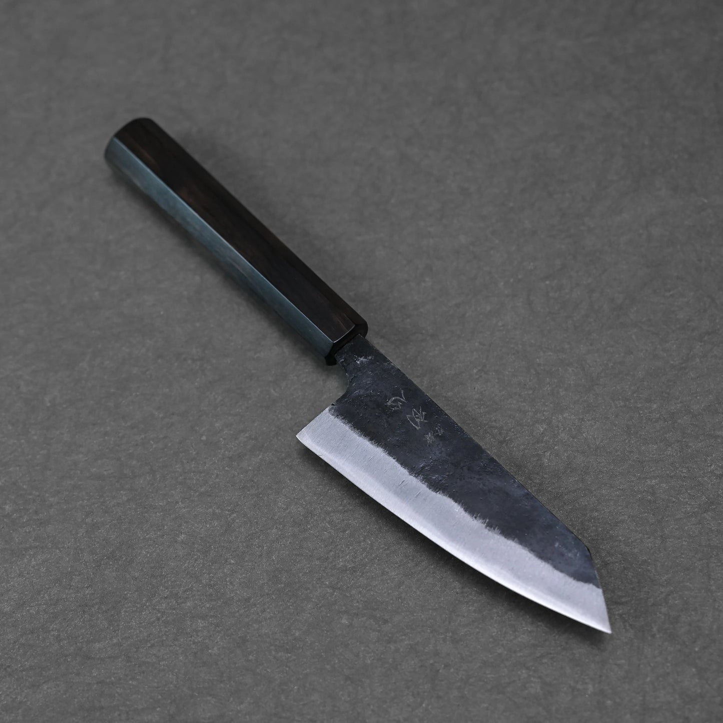 The Hatsukokoro Kurouchi Aogami#2 Ko-Bunka 120mm, a Japanese kitchen knife with a dark wooden handle and short, wide blade, rests on a gray stone surface.