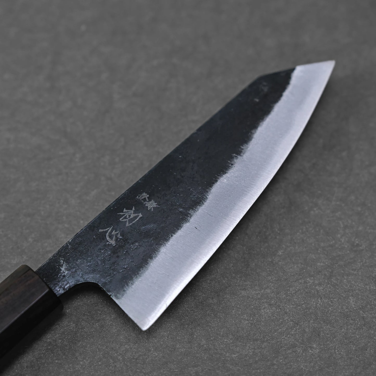 The Hatsukokoro Kurouchi Aogami#2 Ko-Bunka 120mm kitchen knife by Hatsukokoro features a dark handle and blade with a polished edge and kurouchi finish, plus Japanese characters etched near the handle, shown resting on a gray surface.