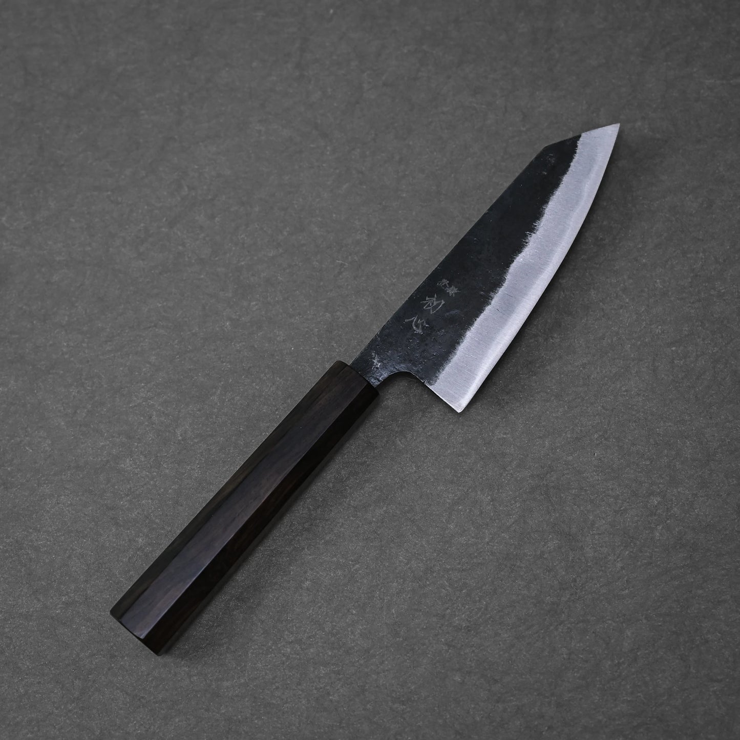 The Hatsukokoro kurouchi aogami#2 ko-bunka 120mm kitchen knife by Hatsukokoro, featuring a dark wooden handle and partially polished Aogami #2 blade, rests on a dark gray textured surface.