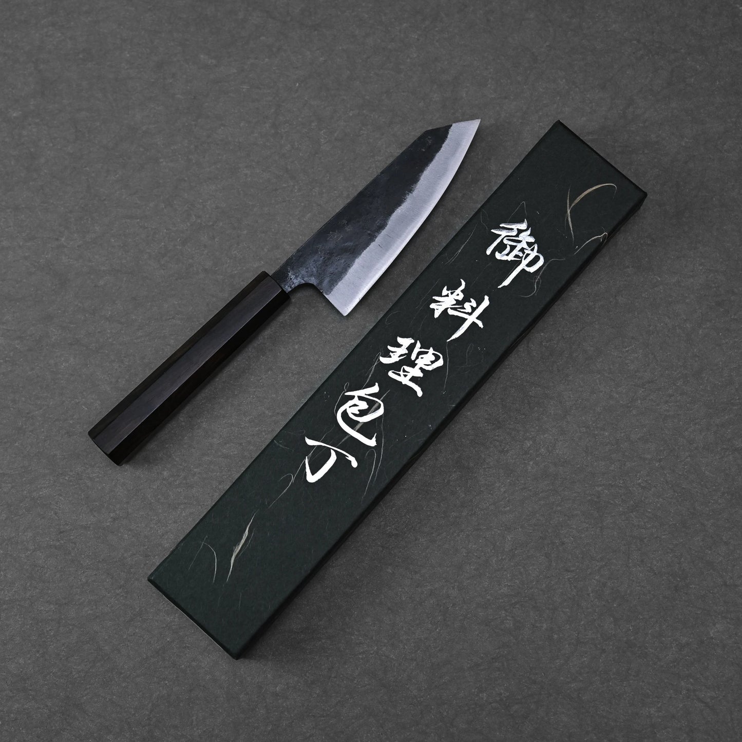 The Hatsukokoro Kurouchi Aogami#2 Ko-Bunka 120mm kitchen knife with a black handle lies next to a black box adorned with white Japanese calligraphy, set on a dark textured background.