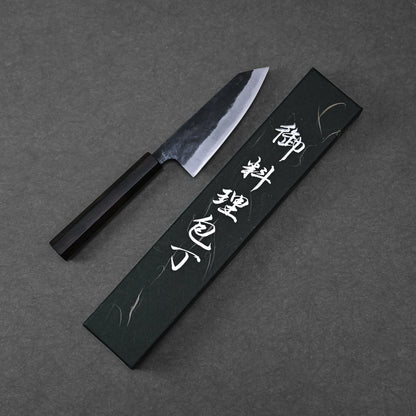 The Hatsukokoro Kurouchi Aogami#2 Ko-Bunka 120mm kitchen knife with a black handle lies next to a black box adorned with white Japanese calligraphy, set on a dark textured background.