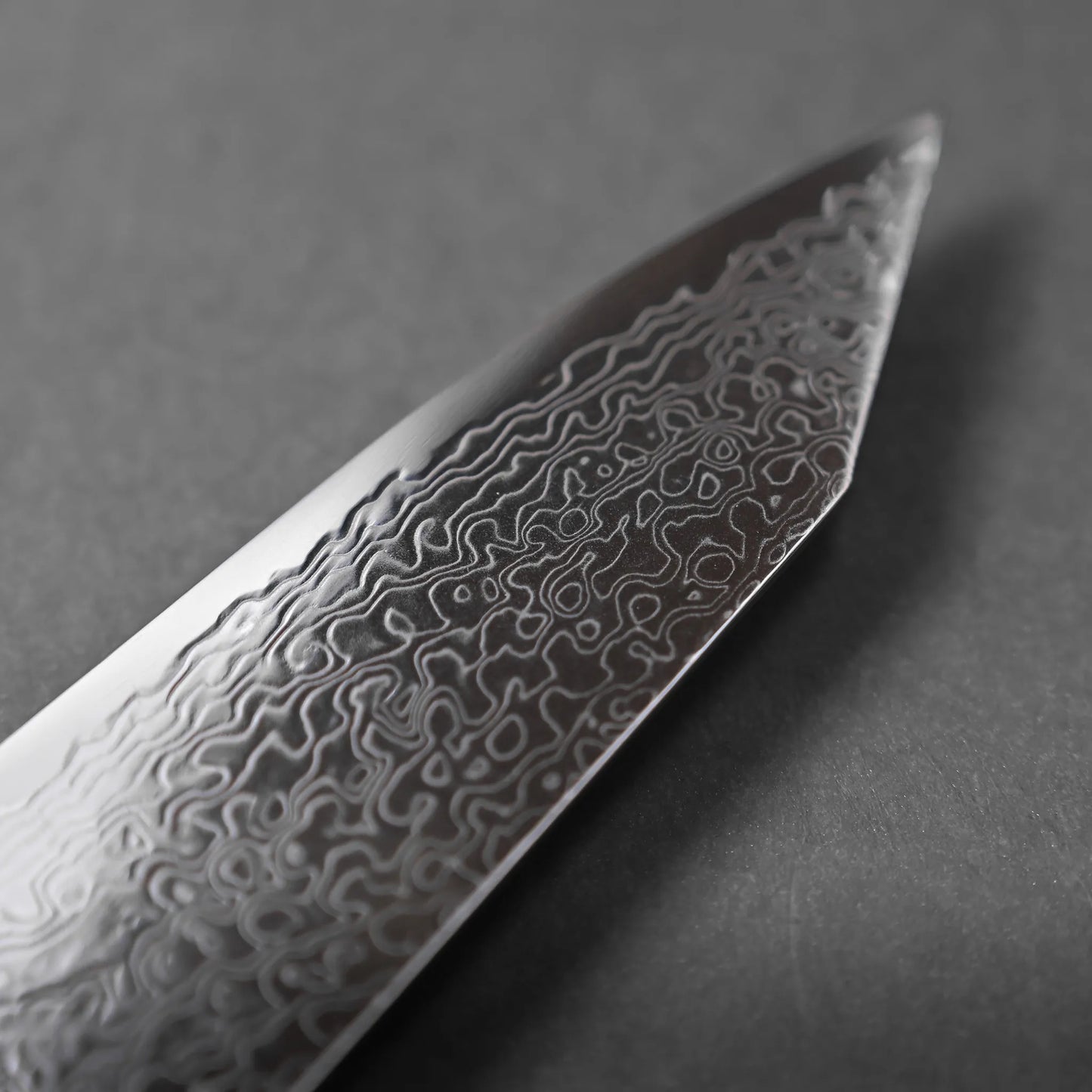 Close-up of the tip of a knife blade featuring visible, intricate Damascus steel patterns, set against a plain, dark gray background, highlighting the unique artistry of Damascus craftsmanship.