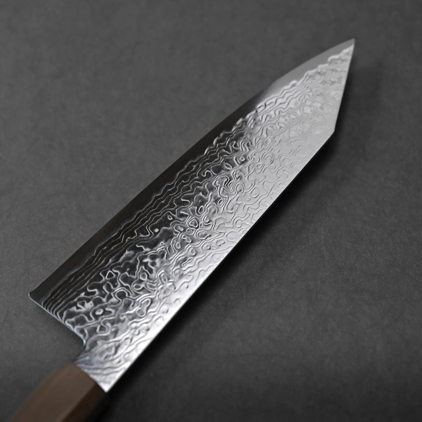 A close-up of a knife with a patterned Damascus steel blade and an elegant wooden handle, placed on a dark gray surface.
