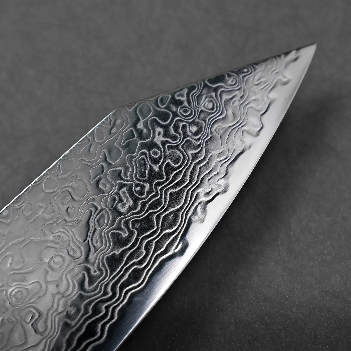Close-up of the tip of a knife blade, featuring intricate wavy patterns characteristic of Damascus steel, set against a plain dark background that highlights its craftsmanship.