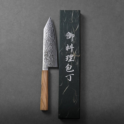 A Japanese-style chef knife with a patterned blade and wooden handle rests next to a sleek black box displaying white Japanese characters, all set against a dark gray background.