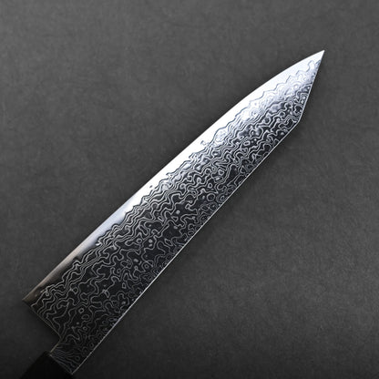 A close-up of a ktip gyuto knife with a patterned Damascus steel blade positioned diagonally on a dark gray background. The intricate lines and waves of the steel are clearly visible.