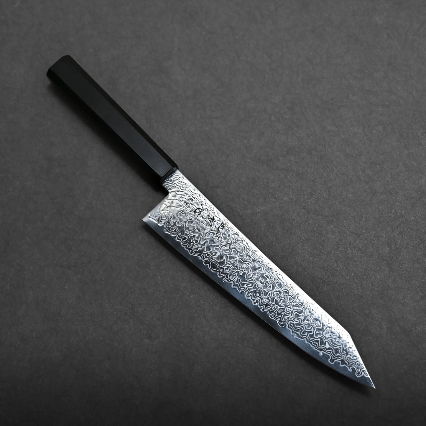 A kiritsuke Japanese chef knife with a black handle and a patterned Damascus steel blade is placed on a dark gray surface.