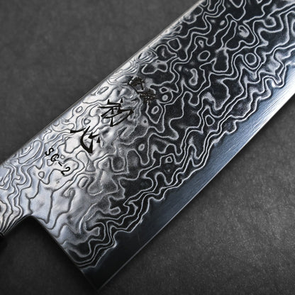 Close-up of a kiritsuke gyuto Japanese chef knife blade, showcasing a detailed, wavy Damascus steel pattern and engraved characters near the edge, set against a dark background.