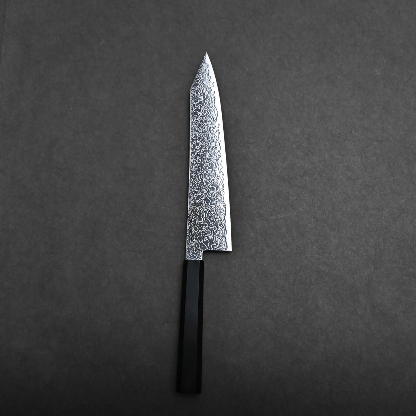 A single kiritsuke Japanese chef knife with a patterned blade and a black rectangular handle is placed on a dark, plain surface.