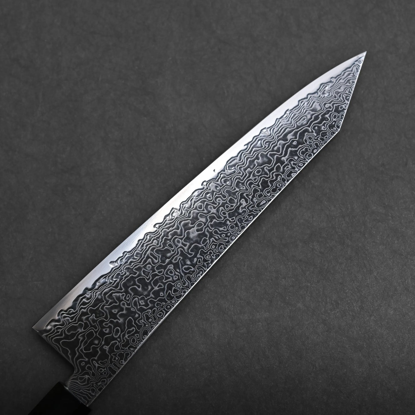 A close-up of a ktip gyuto chef’s knife with a patterned Damascus steel blade, showing intricate wavy designs, set against a dark textured background.