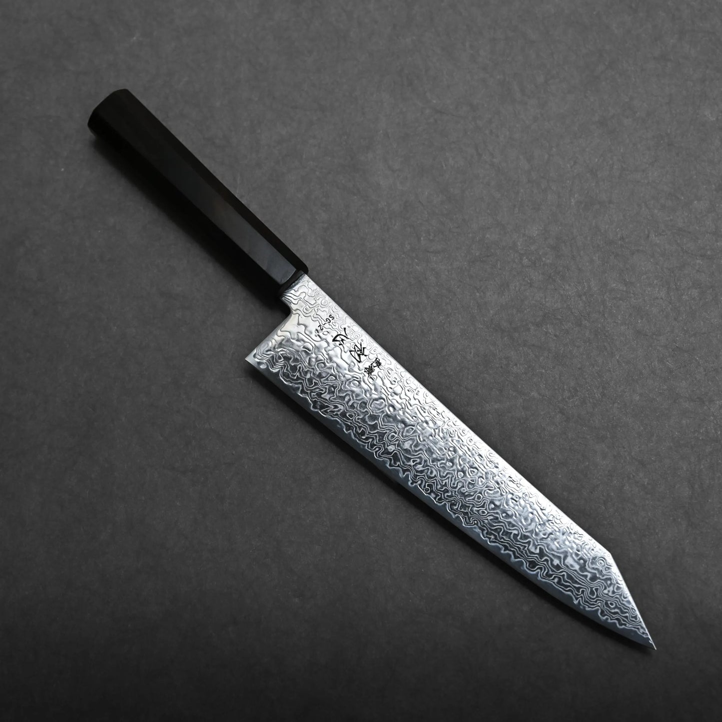 A kiritsuke gyuto knife with a dark handle and a patterned Damascus steel blade rests on a dark gray textured surface. The blade features intricate wavy designs and Japanese characters etched near the handle.