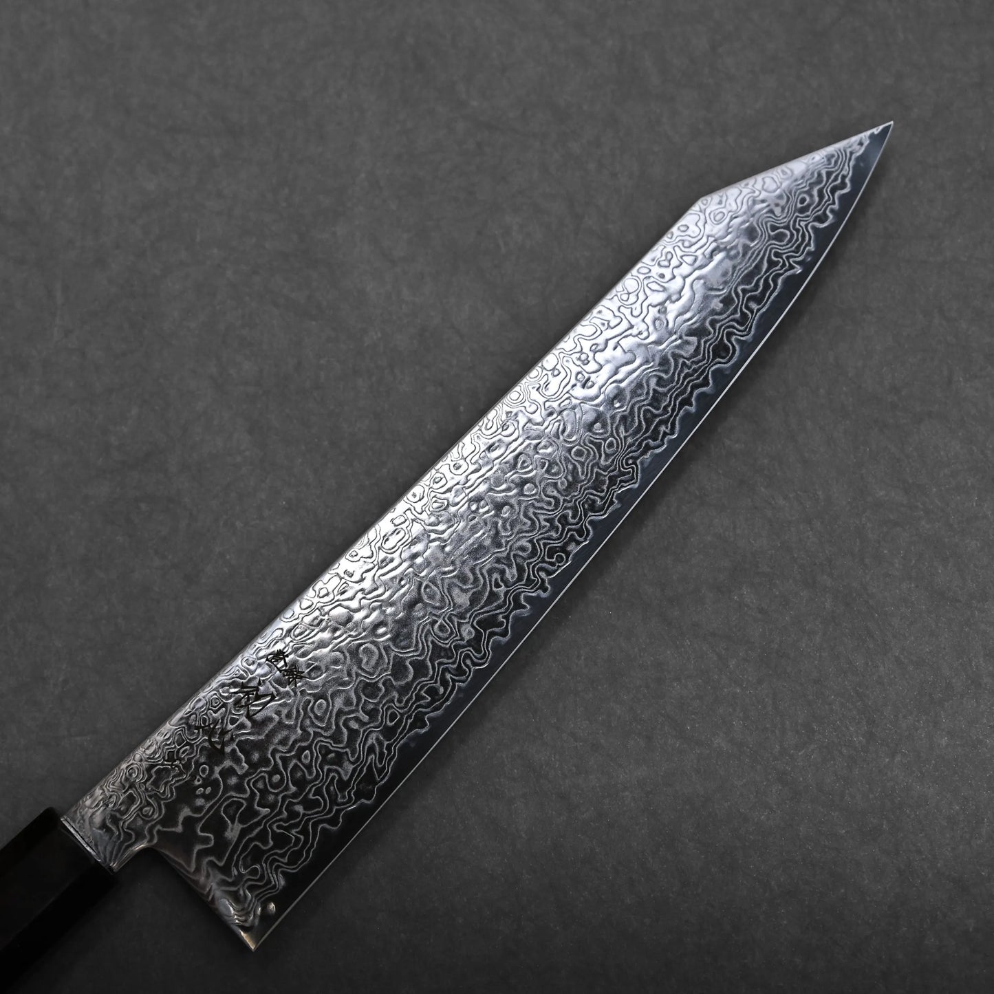 A close-up of a kiritsuke Japanese chef knife with a patterned Damascus steel blade, featuring intricate swirling designs, resting on a dark gray textured surface.