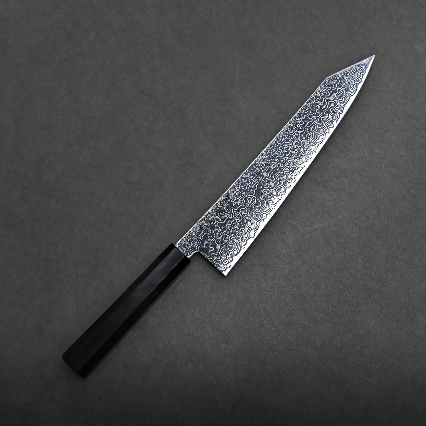 A Hatsukokoro Ryuhyo SG2 damascus kiritsuke gyuto 240mm with a patterned Damascus steel blade and a dark, angular handle, placed on a dark gray background.